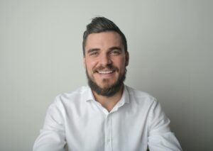 Portrait of a young bearded man smiling, wearing a white shirt indoors against a gray background.